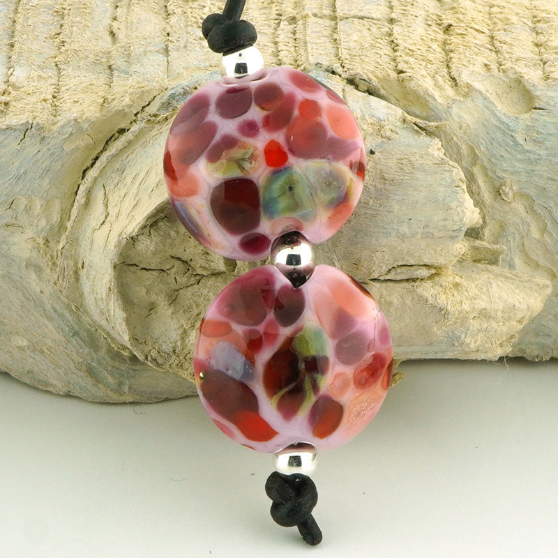 A close-up of a handmade lampara bead necklace featuring two round, patterned pink and red beads with black cord and silver spacer beads, set against a textured light gray stone background.