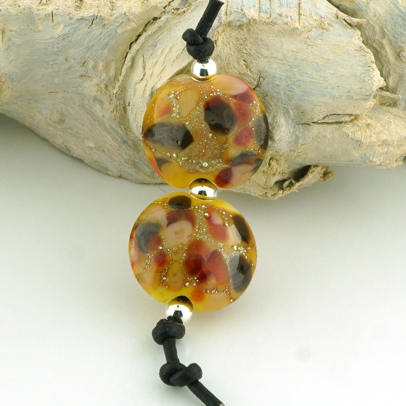 Two lentil shaped glass lampwork beads with a marbled pattern of amber, red, brown, and black, suspended on a black cord with silver spacers, displayed against a piece of driftwood.
