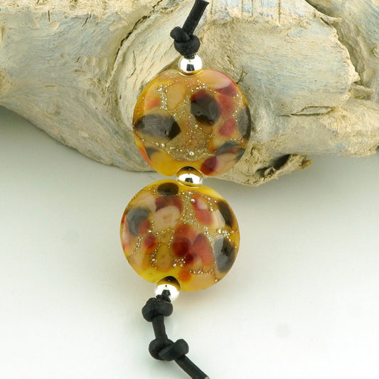Two lentil shaped glass lampwork beads with a marbled pattern of amber, red, brown, and black, suspended on a black cord with silver spacers, displayed against a piece of driftwood.