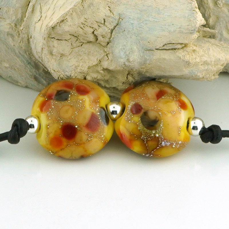 Two lentil shaped glass lampwork beads with a marbled pattern of amber, red, brown, and black, suspended on a black cord with silver spacers, displayed against a piece of driftwood.
