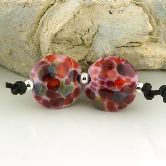 Two round glass lampwork beads with a mottled pattern of red, purple, orange, and green on a white cord, displayed against a rustic light gray wood background.