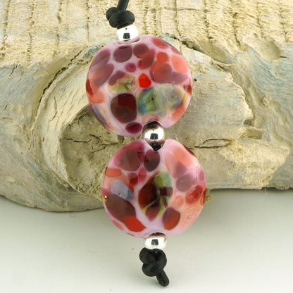A close-up of a handmade lampara bead necklace featuring two round, patterned pink and red beads with black cord and silver spacer beads, set against a textured light gray stone background.