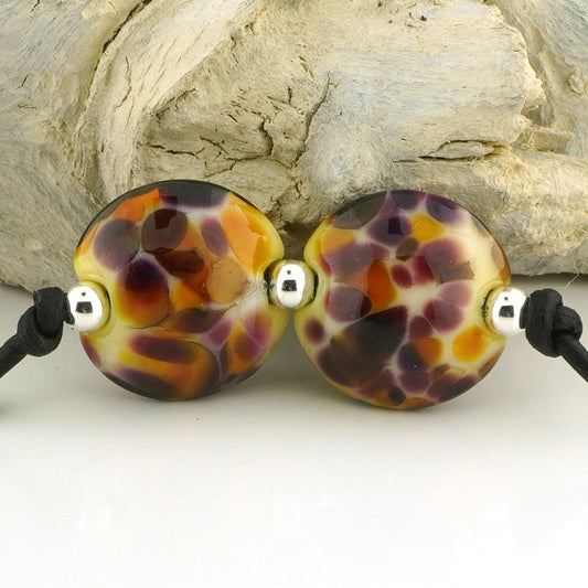 Two round glass lampwork beads with a marbled pattern of purple, orange, yellow, and black, strung on black cords with small silver spacer beads, displayed against a light gray background.