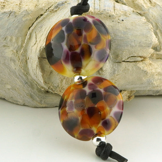 Two round glass lampwork beads with a marbled pattern of orange, brown, purple, and cream, strung on a black cord with silver spacer beads, set against a rustic white wood background.