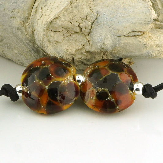Two round glass lampwork beads with a mottled pattern of dark brown, amber orange, and black, separated by small silver spacer beads on a black cord, displayed against a light gray textured background.