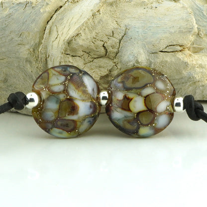 Two round glass beads with a marbled pattern of brown, cream, and light blue, threaded on a black cord with silver spacers, displayed against a textured light wood background.