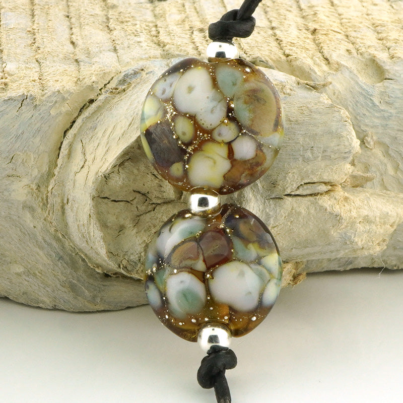 Two round glass beads with a marbled pattern of brown, cream, and light blue, threaded on a black cord with silver spacers, displayed against a textured light wood background.