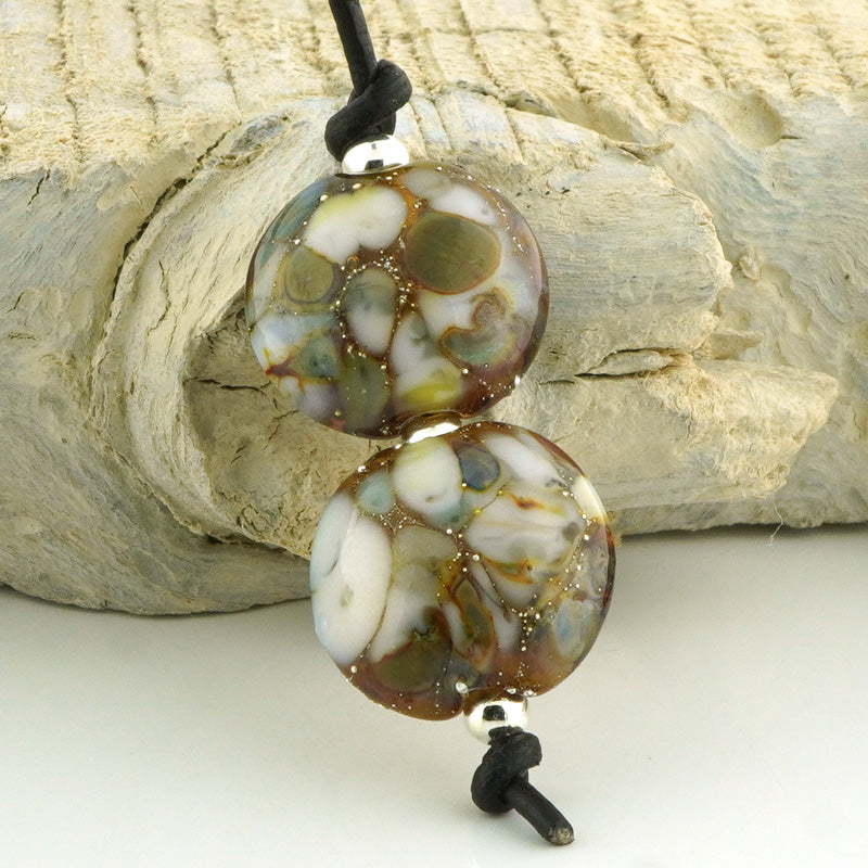 Two round handmade lampwork beads with a marbled pattern of white, brown, green, and gold, threaded on a black cord, displayed against a rustic wood background.