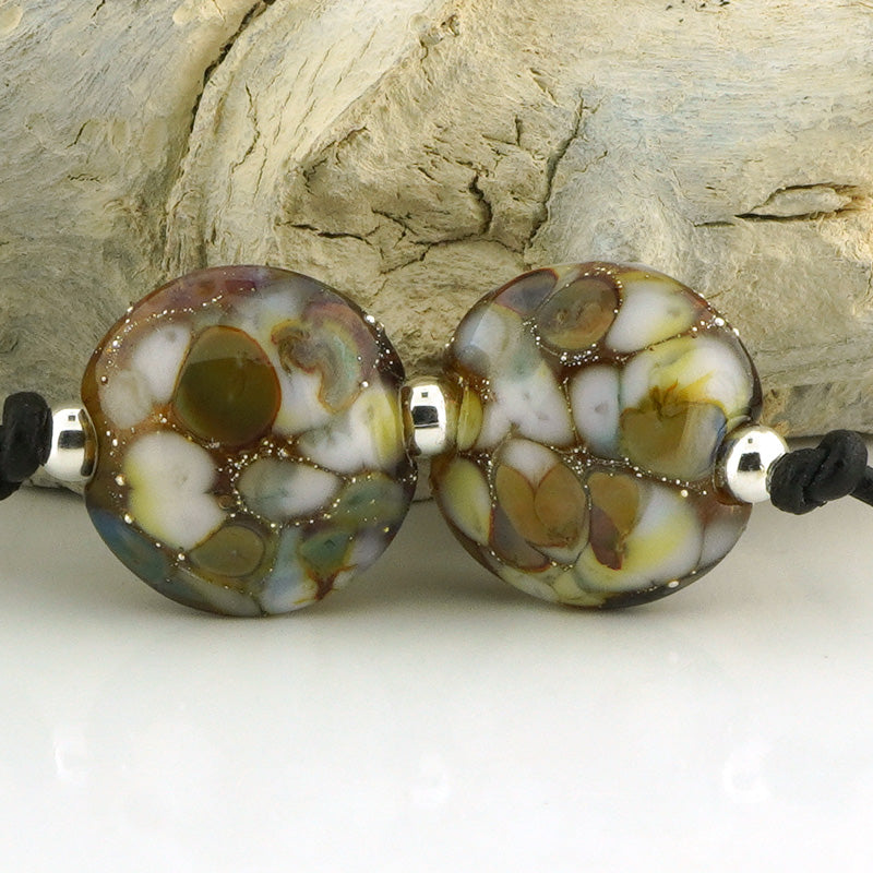 Two round glass lampwork beads with a marbled pattern of white, brown, olive green, and gray, separated by small silver metal spacers and threaded onto a black cord, set against a background of textured driftwood.