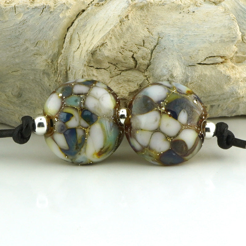 Two round handmade lampwork beads with a marbled pattern of white, blue, green, and brown, separated by small silver spacer beads on a black cord, set against a background of light-colored driftwood.