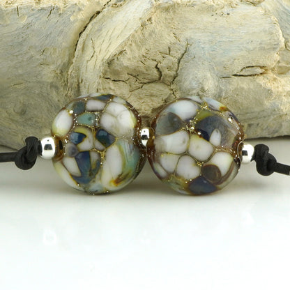 Two round handmade lampwork beads with a marbled pattern of white, blue, green, and brown, separated by small silver spacer beads on a black cord, set against a background of light-colored driftwood.