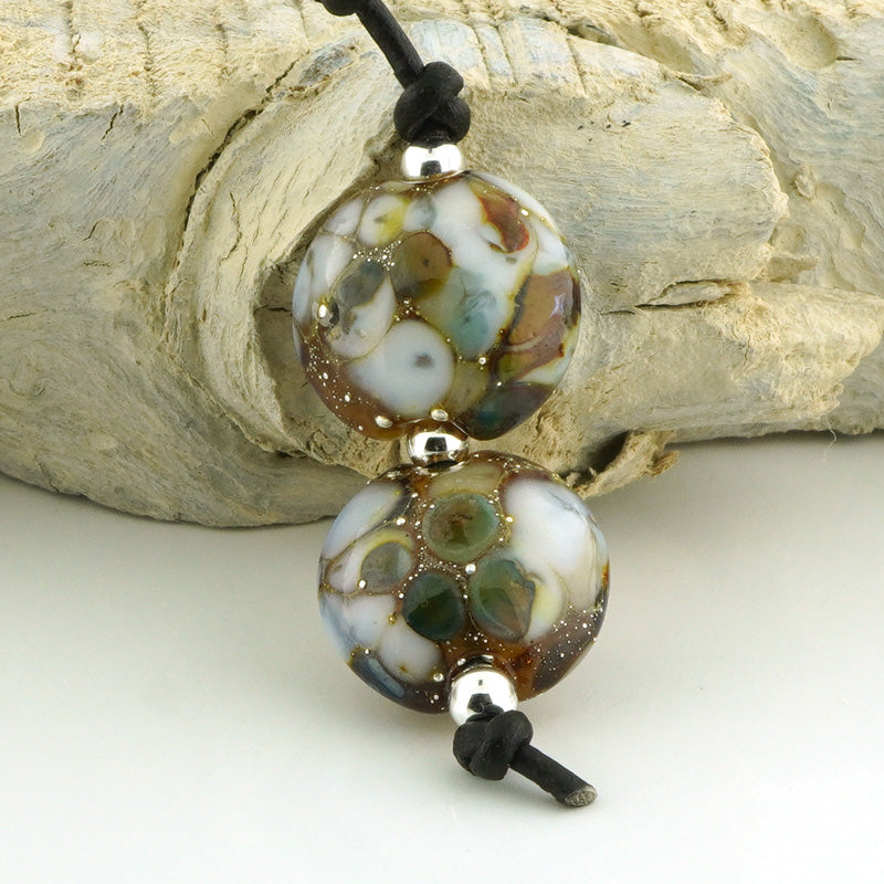 Two round handmade lampwork beads with a marbled pattern of white, brown, green, and gold, threaded on a black cord, displayed against a rustic wood background.
