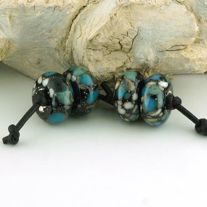 Three round glass lampwork beads with a marbled pattern of turquoise blue, black, and white, threaded on a black cord, displayed against a light-colored wood background.